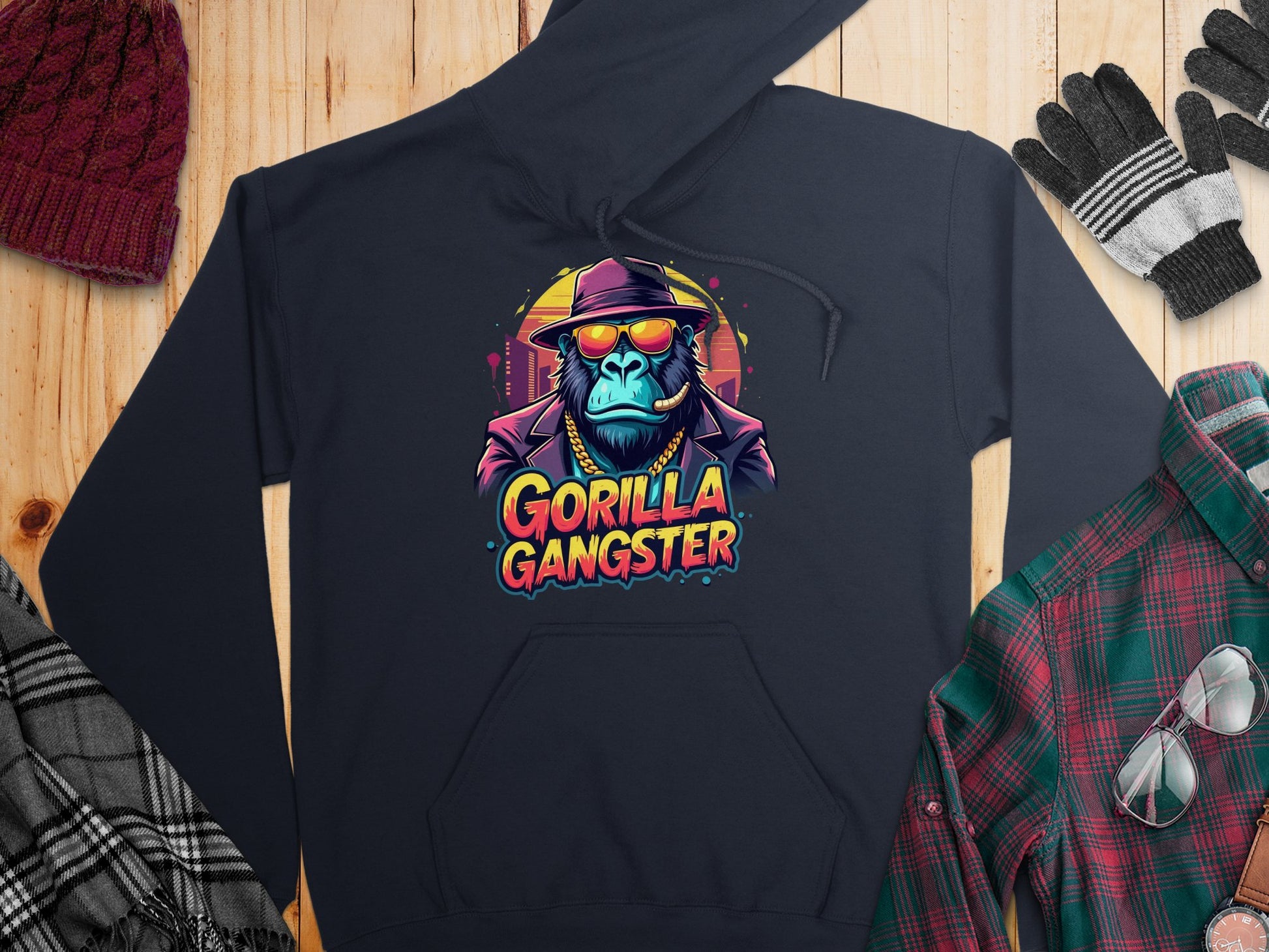 A dark hoodie from Garment Graphics features a striking gorilla in a hat and sunglasses with Gorilla Gangster printed below, epitomizing streetwear. Enhanced by a vibrant cityscape backdrop, winter clothing items are stylishly displayed on a wooden surface.