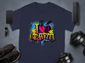 A dark gray T-shirt from Garment Graphics displays a vibrant I Love Graffiti design with a broken heart, paint splatters, and brick elements. Nearby are a smartphone, smartwatch, black shoes, a speaker, and adjustable dumbbells.