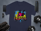 A dark gray T-shirt from Garment Graphics displays a vibrant I Love Graffiti design with a broken heart, paint splatters, and brick elements. Nearby are a smartphone, smartwatch, black shoes, a speaker, and adjustable dumbbells.