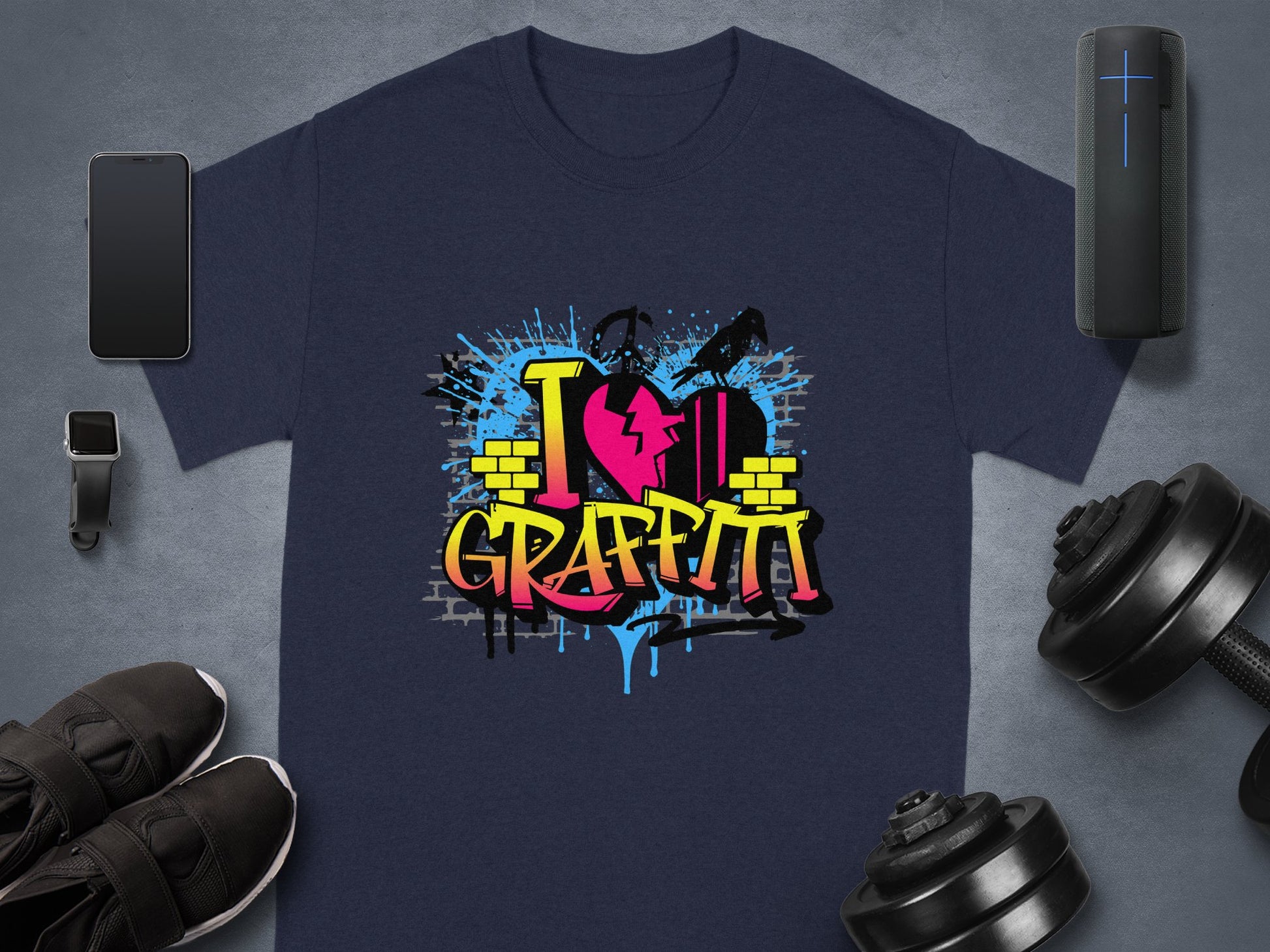 A dark gray T-shirt from Garment Graphics displays a vibrant I Love Graffiti design with a broken heart, paint splatters, and brick elements. Nearby are a smartphone, smartwatch, black shoes, a speaker, and adjustable dumbbells.