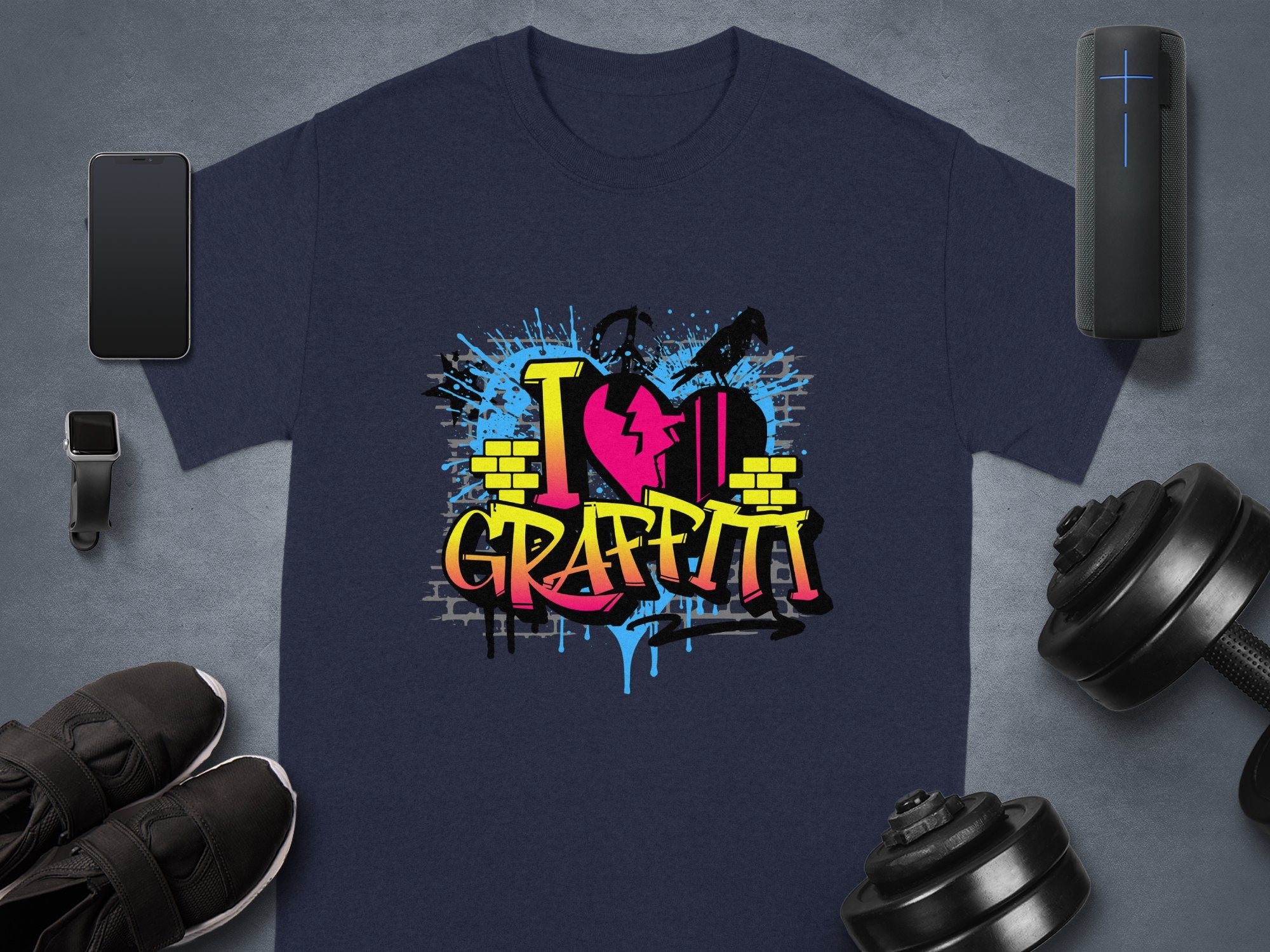 A dark gray T-shirt from Garment Graphics displays a vibrant I Love Graffiti design with a broken heart, paint splatters, and brick elements. Nearby are a smartphone, smartwatch, black shoes, a speaker, and adjustable dumbbells.