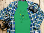 A classic fit green t-shirt by Royal Mail - 24 Tracked, featuring Lucky Just Blessed Shamrock and shamrock designs, rests on a checkered shirt. Nearby are black headphones, a camera, and a to-go coffee cup on the wooden surface.