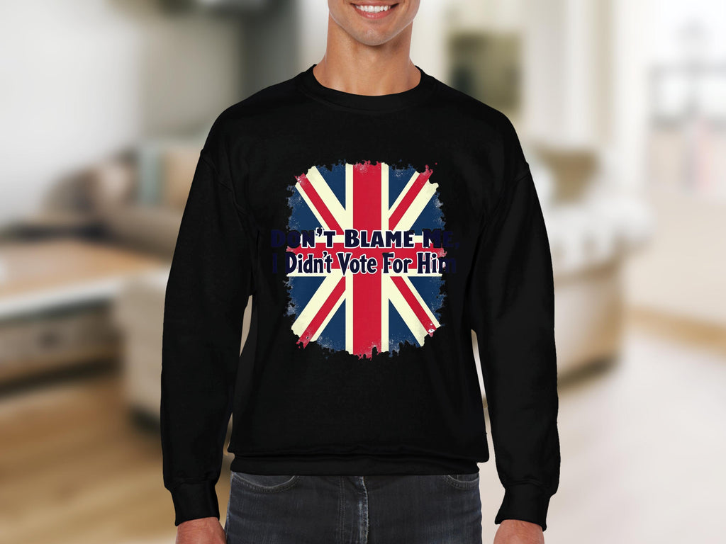A person models a comfy black sweatshirt from Garment Graphics, featuring a bold distressed Union Jack flag design and the text DONT BLAME ME, I didnt vote for him, set against a blurred, light-colored room.
