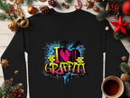 A black Garment Graphics sweatshirt with I ♥ Graffiti in bold, urban design rests on a wooden surface. Encircled by pine cones, ornaments, and a coffee cup, it exudes a festive holiday vibe.