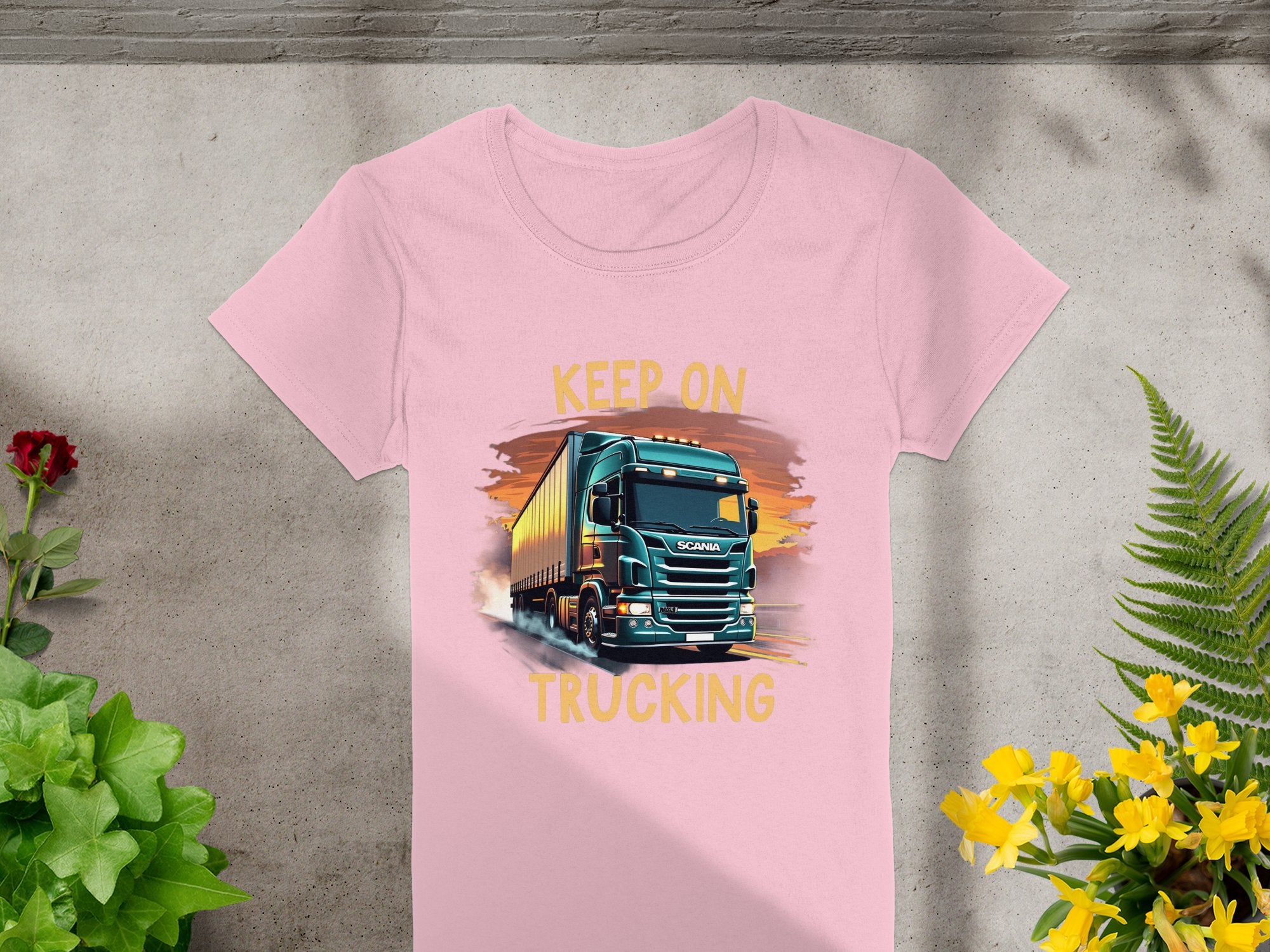 This Garment Graphics pink T-shirt showcases a bold black semi-truck driving at sunset with KEEP ON TRUCKING around the graphic, ideal for trucking fans. Photographed on concrete surrounded by greenery, it captures the essence of Keep on Trucking T-shirts.