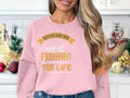 A smiling woman with long hair stands in a festively decorated kitchen, wearing a pink sweatshirt from Garment Graphics, featuring the text Father and Son Fishing, Partners for Life and an illustration of a boat with fish and hooks.