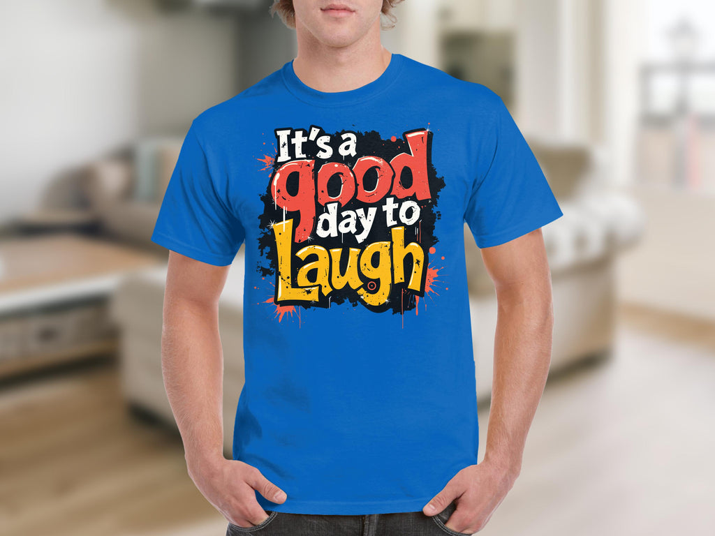 Someone is wearing a vibrant Its a Good Day to Laugh t-shirt from Garment Graphics, featuring cheerful red, black, white, and yellow stylized letters. The background is a blurred indoor setting.