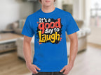 Someone is wearing a vibrant Its a Good Day to Laugh t-shirt from Garment Graphics, featuring cheerful red, black, white, and yellow stylized letters. The background is a blurred indoor setting.