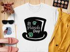 A St Patricks Day white sweatshirt with a top hat and shamrocks by Garment Graphics is laid on a wooden surface, surrounded by a yellow jacket, pink shoes, cosmetics, makeup brushes, a blue pouch, sunglasses, and nail polish.