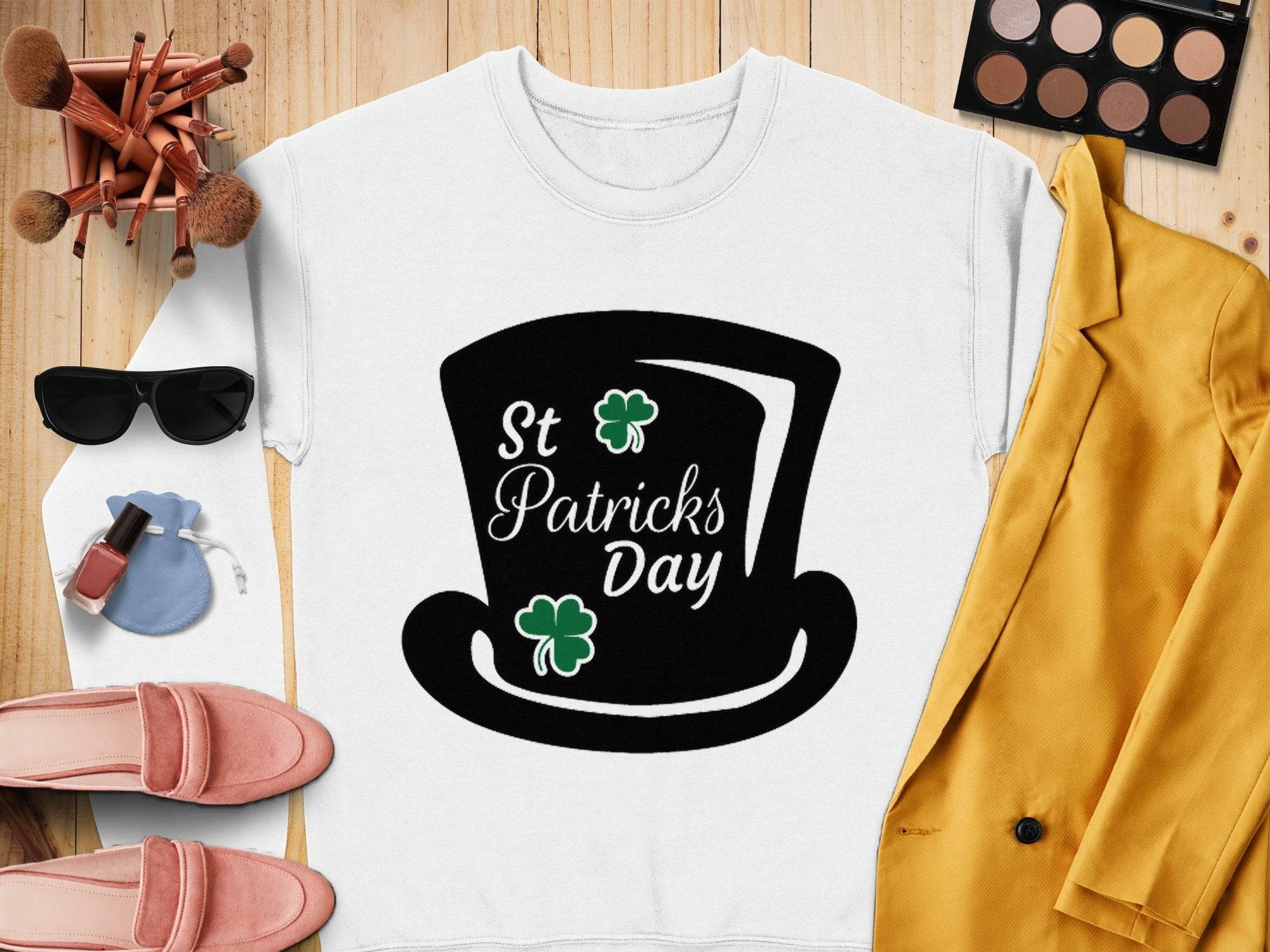 A St Patricks Day white sweatshirt with a top hat and shamrocks by Garment Graphics is laid on a wooden surface, surrounded by a yellow jacket, pink shoes, cosmetics, makeup brushes, a blue pouch, sunglasses, and nail polish.