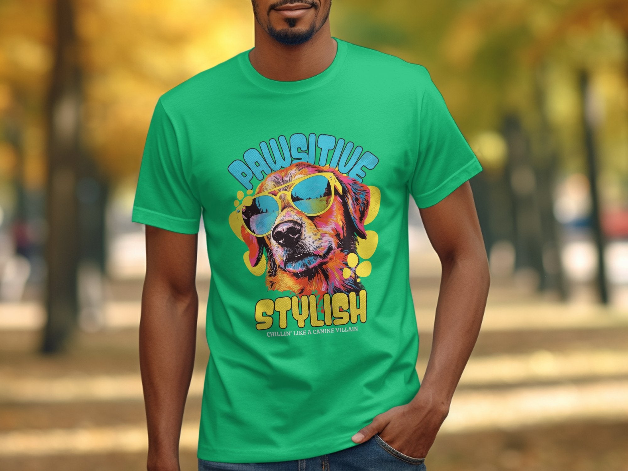 Wearing a bright green Garment Graphics T-shirt adorned with a colorful dog in sunglasses, the shirt reads Paw-sitive Stylish and Chillin Like A Canine Villain, matching the serene vibe of the autumn trees in the background.