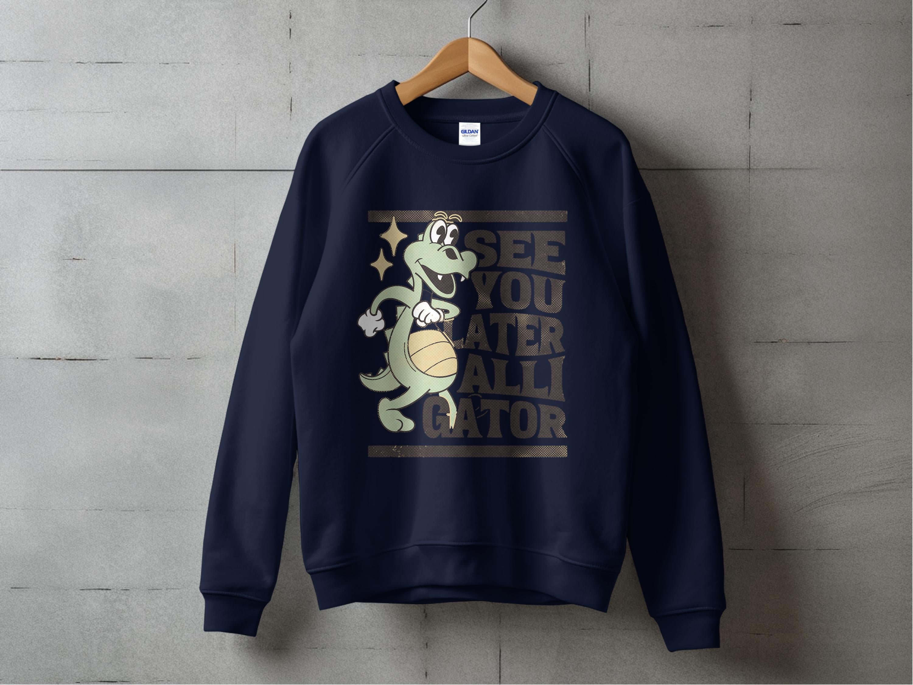 Garment Graphics navy blue unisex sweatshirt showcases a cartoon alligator with a beach ball and the caption, See you later alligator on the front. Displayed against a concrete wall backdrop.