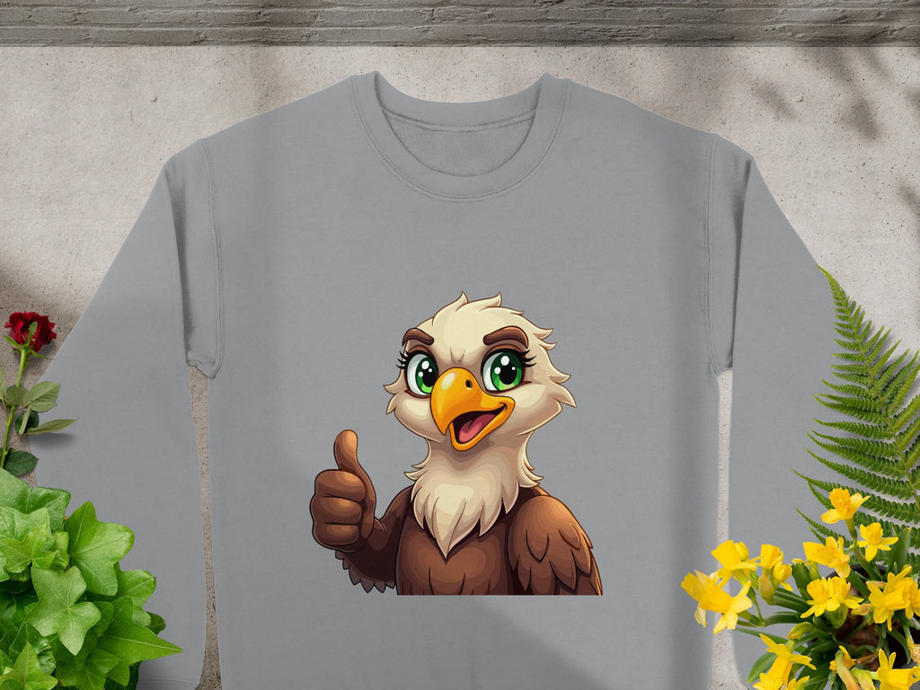 This cheerful cartoon eagle sweatshirt by Garment Graphics features a gray color, an eagle with glowing eyes giving a thumbs-up, set against lush green plants and small yellow flowers. Unisex sizing ensures style and comfort for all.