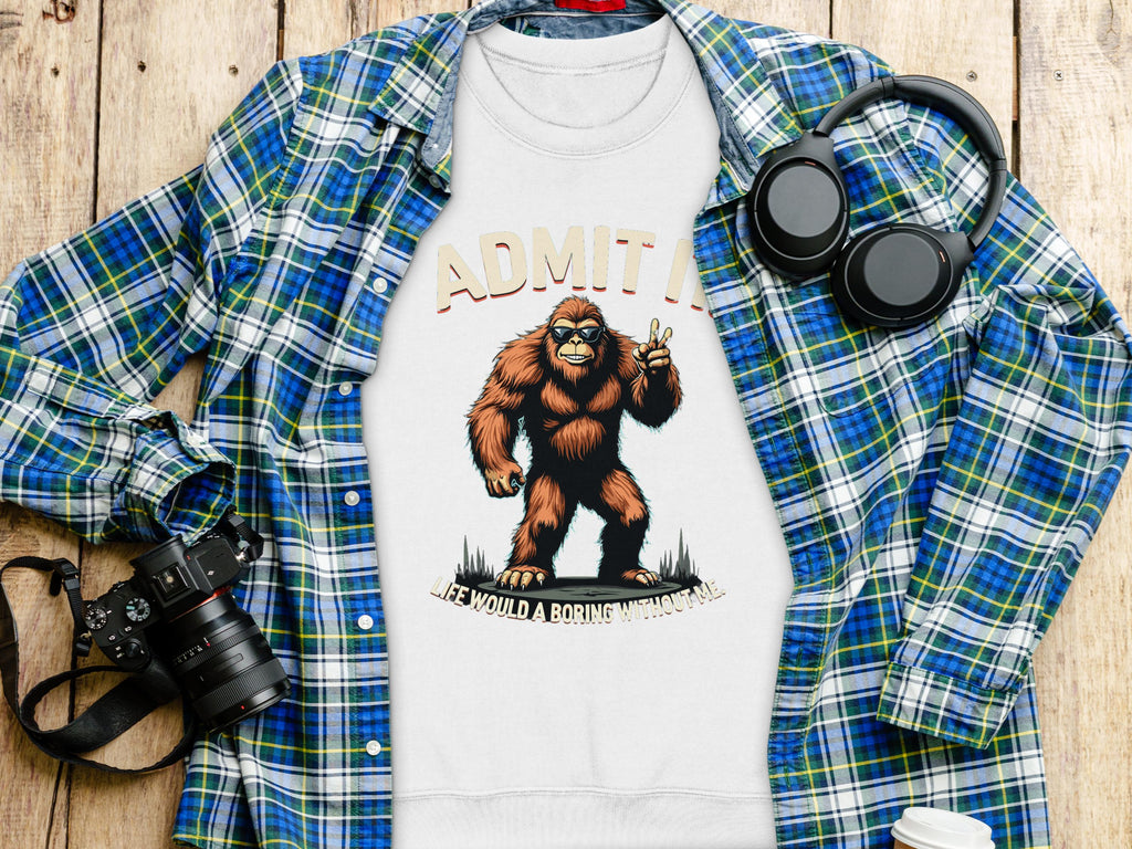 A plaid shirt, a Garment Graphics T-shirt with Bigfoot and Admit it, life would be boring without me, headphones, a camera, and a coffee cup are arranged on a wooden surface.