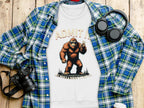 A plaid shirt, a Garment Graphics T-shirt with Bigfoot and Admit it, life would be boring without me, headphones, a camera, and a coffee cup are arranged on a wooden surface.