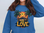 A person in a blue Garment Graphics sweatshirt, featuring graphic palm trees, a sunset over mountains, and a flying bird. The bold One Love print pairs perfectly with their dark brown wavy hair.