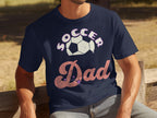 A dad sits outdoors on a wooden bench in a Garment Graphics navy blue T-shirt, featuring Soccer Dad with a soccer ball. The backdrop is a grassy landscape.