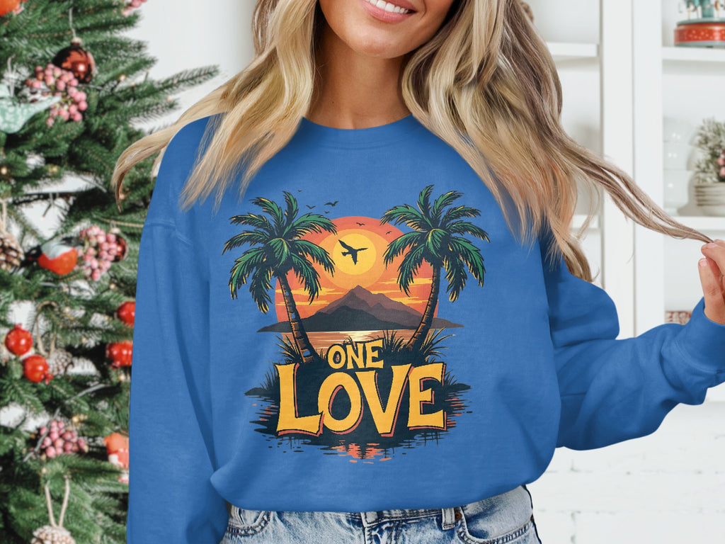 Indoors by a decorated Christmas tree, a person wears a stylish Garment Graphics sweatshirt. The blue top features a tropical design with palm trees, a stunning One Love sunset, and the words One Love.