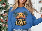 Indoors by a decorated Christmas tree, a person wears a stylish Garment Graphics sweatshirt. The blue top features a tropical design with palm trees, a stunning One Love sunset, and the words One Love.