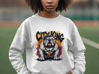 A person wearing a Garment Graphics Bulldog Sweatshirt, showcasing a bulldog with a gold chain and the phrase City King above it, stands against a blurred outdoor background.