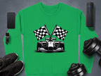 Garment Graphics green sweatshirt showcases a black and white race car with crossed checkered flags. The scene includes a smartphone, smartwatch, wireless speaker, dumbbells, and black sneakers on a gray background.