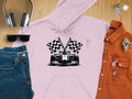 A flat lay features a Garment Graphics light pink hoodie with a bold racing theme, including a race car and checkered flag graphic. Surrounding items include jeans, sunglasses, headphones, keys, an orange shirt, and a gold watch on a wooden surface.