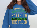 A person wears a Garment Graphics blue sweatshirt with a bold, humorous design: If an Englishman gets run down by a truck, he apologizes to the truck. The varying text colors—green, beige, black—add playfulness. Available in unisex sizing for everyone.