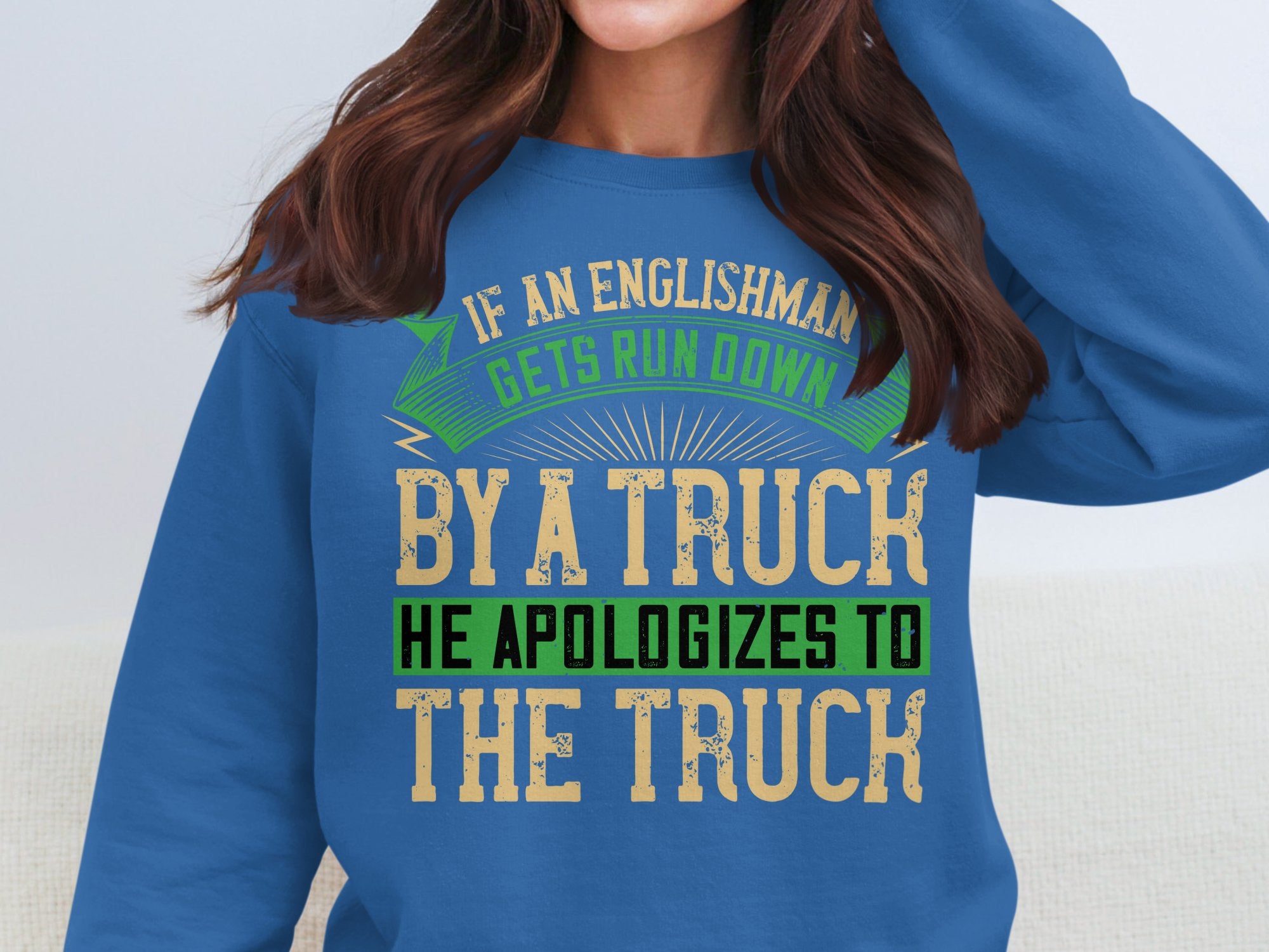 A person wears a Garment Graphics blue sweatshirt with a bold, humorous design: If an Englishman gets run down by a truck, he apologizes to the truck. The varying text colors—green, beige, black—add playfulness. Available in unisex sizing for everyone.