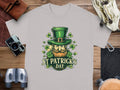 Garment Graphics offers a gray T-shirt with St. Patricks Day design featuring Irish pride—a smiling bearded face, green hat, and shamrocks. The shirt reads St. Patricks Day, pictured with a camera, binoculars, and leather pouches on wood surface—ideal for celebrating in style.