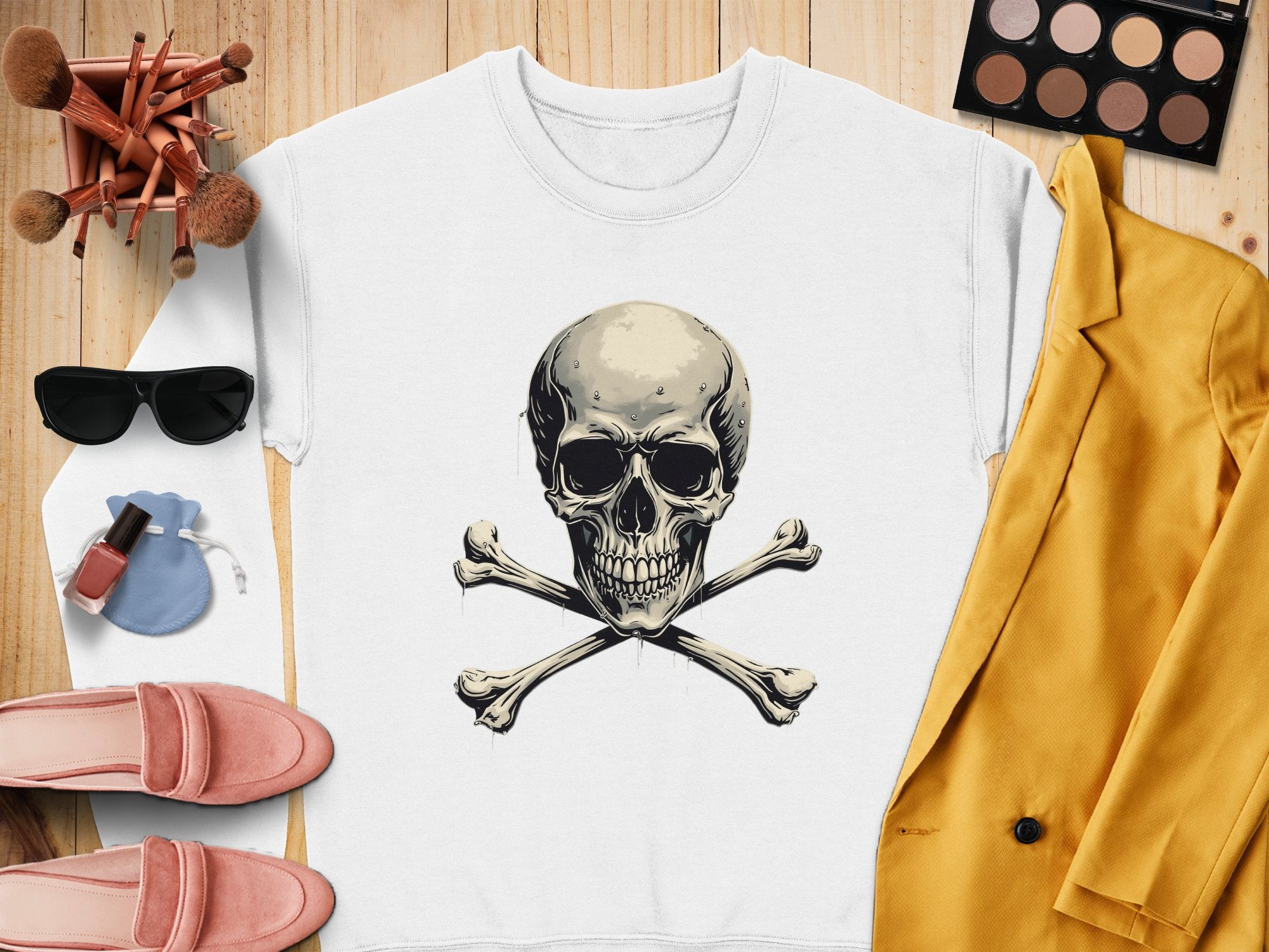 On a wooden surface, a white Garment Graphics sweatshirt with a skull and crossbones design sets an edgy scene, accented by makeup brushes, sunglasses, a makeup palette, a yellow jacket, pink shoes, nail polish, and a small blue pouch.