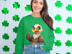 A woman stands against a wall covered in cheerful green shamrocks, wearing Garment Graphics green sweatshirt with a cartoon eagle giving a thumbs-up, paired with denim jeans. She smiles at the camera.