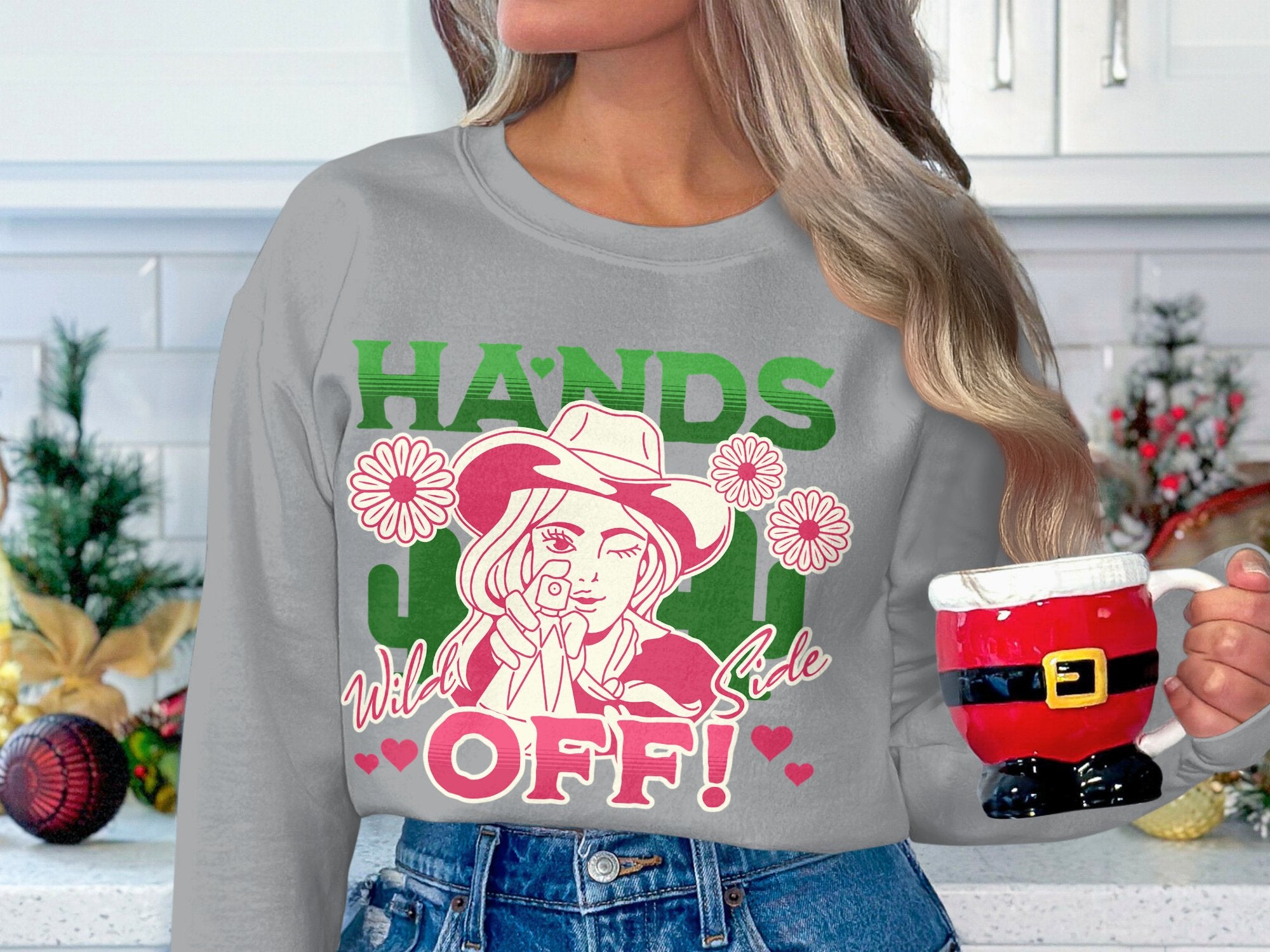 In a kitchen, someone holds a red Santa-themed mug. They are donning the Garment Graphics Hands Off Wild Side Sweatshirt, noted for its loose fit and eye-catching design with a hat-wearing figure, flowers, and vibrant green and pink text.