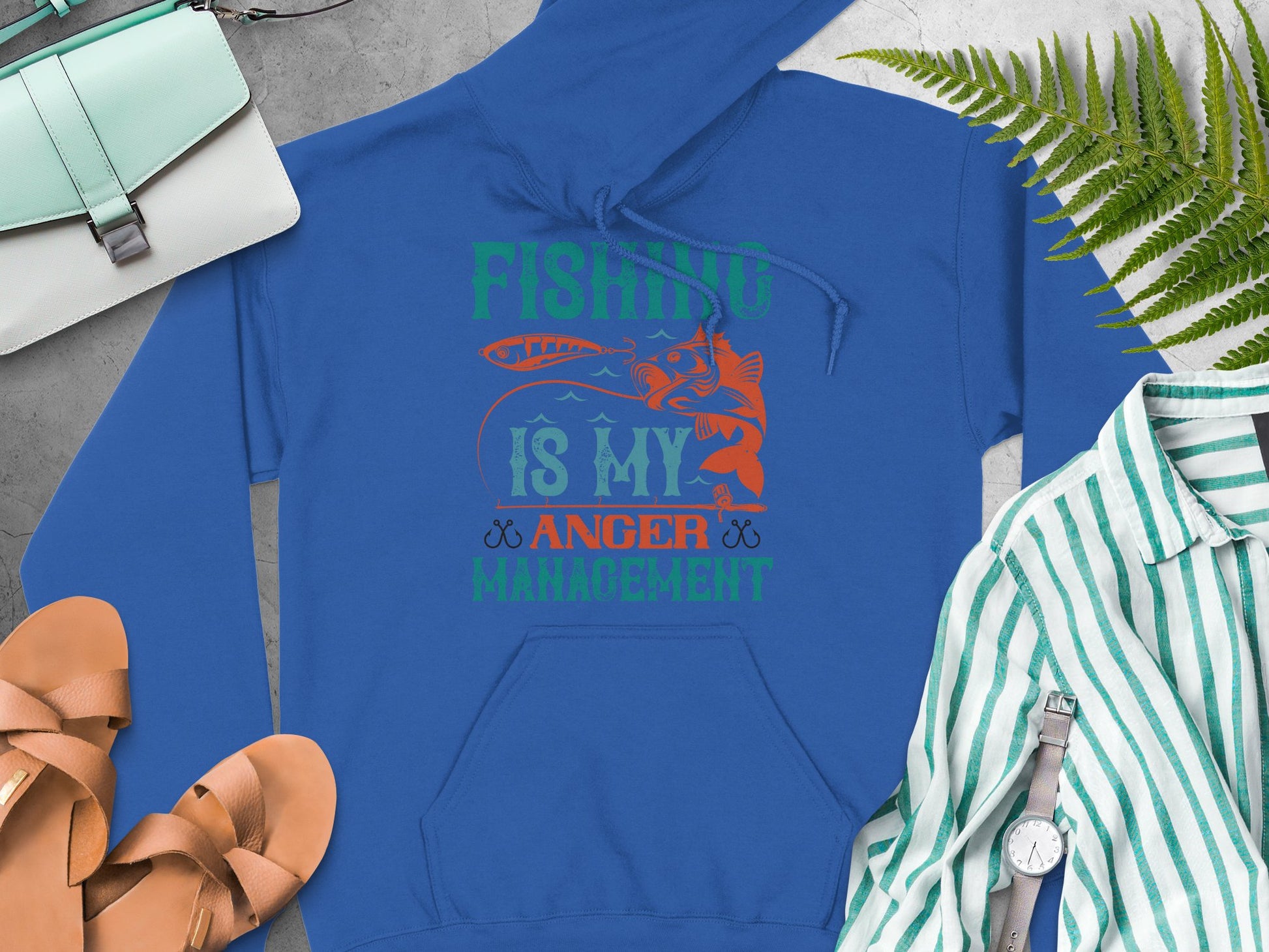 A blue Garment Graphics hoodie with Fishing is my anger management text anchors this stylish ensemble. Laid on concrete, its paired with tan sandals, a striped shirt, a watch, a fern leaf, and a light blue bag for the perfect outdoor look.