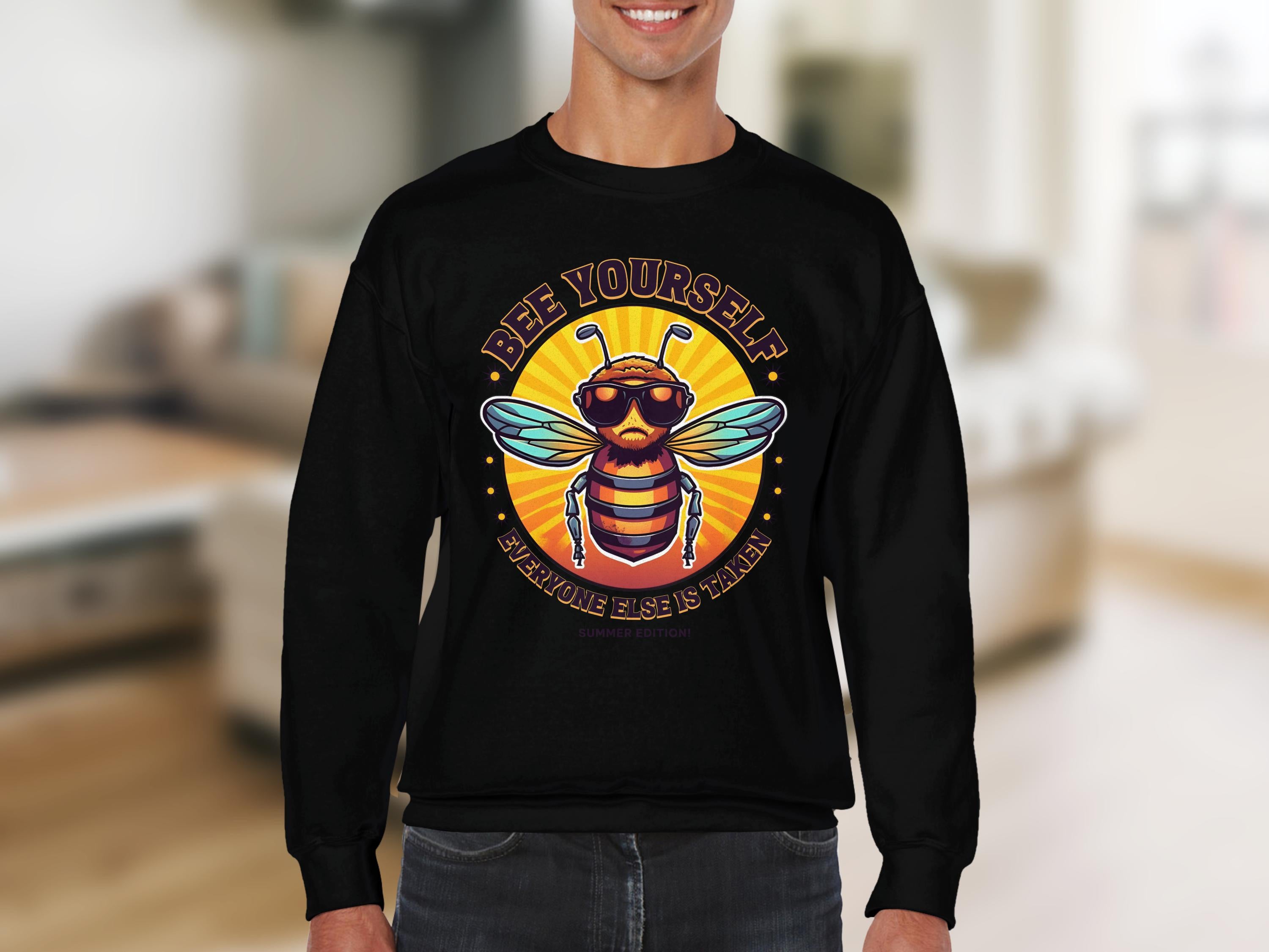 Someone is wearing a black Garment Graphics sweatshirt from the Summer Edition Sweatshirts collection, featuring a vibrant bee in sunglasses with the text Bee Yourself, Everyone Else is Taken against a blurred kitchen or living area backdrop.
