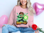 A woman with wavy blond hair wears a Garment Graphics pink sweatshirt featuring animated superhero graphics and the words Its Hero Time! Sitting cross-legged in casual ripped jeans, she holds a red rose beside pink heart-shaped balloons.