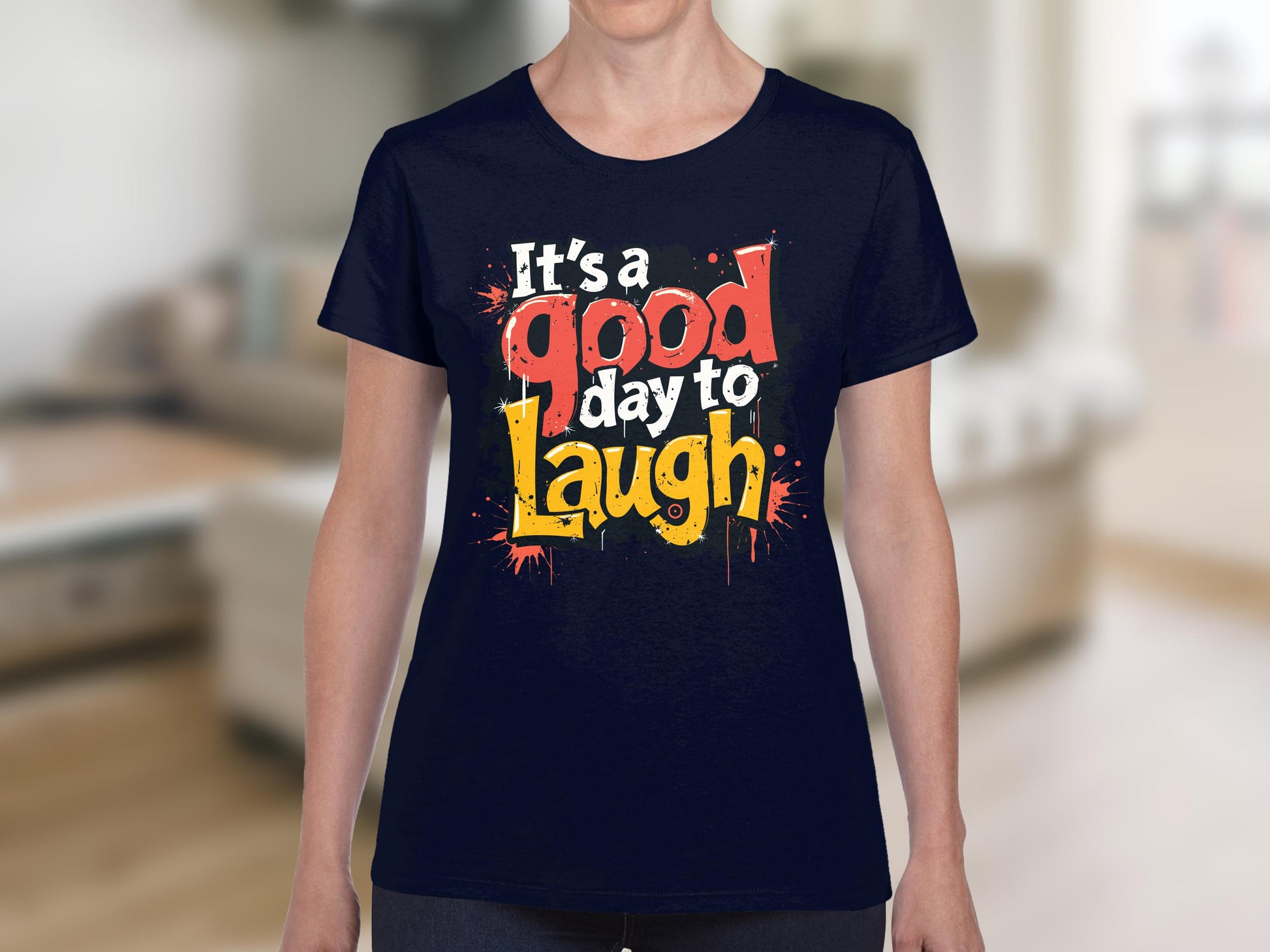 Wearing a semi-fitted T-shirt by Garment Graphics, adorned with bold, colorful text Its a good day to laugh, someone stands in front of light-colored furniture in a blurred interior space.