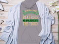 A gray hoodie from Garment Graphics features a humorous quote: If an Englishman gets run over by a truck, he apologizes to the truck in green, yellow, and white letters. The funny design is displayed under a light gray cardigan with decorations, capturing witty English etiquette.