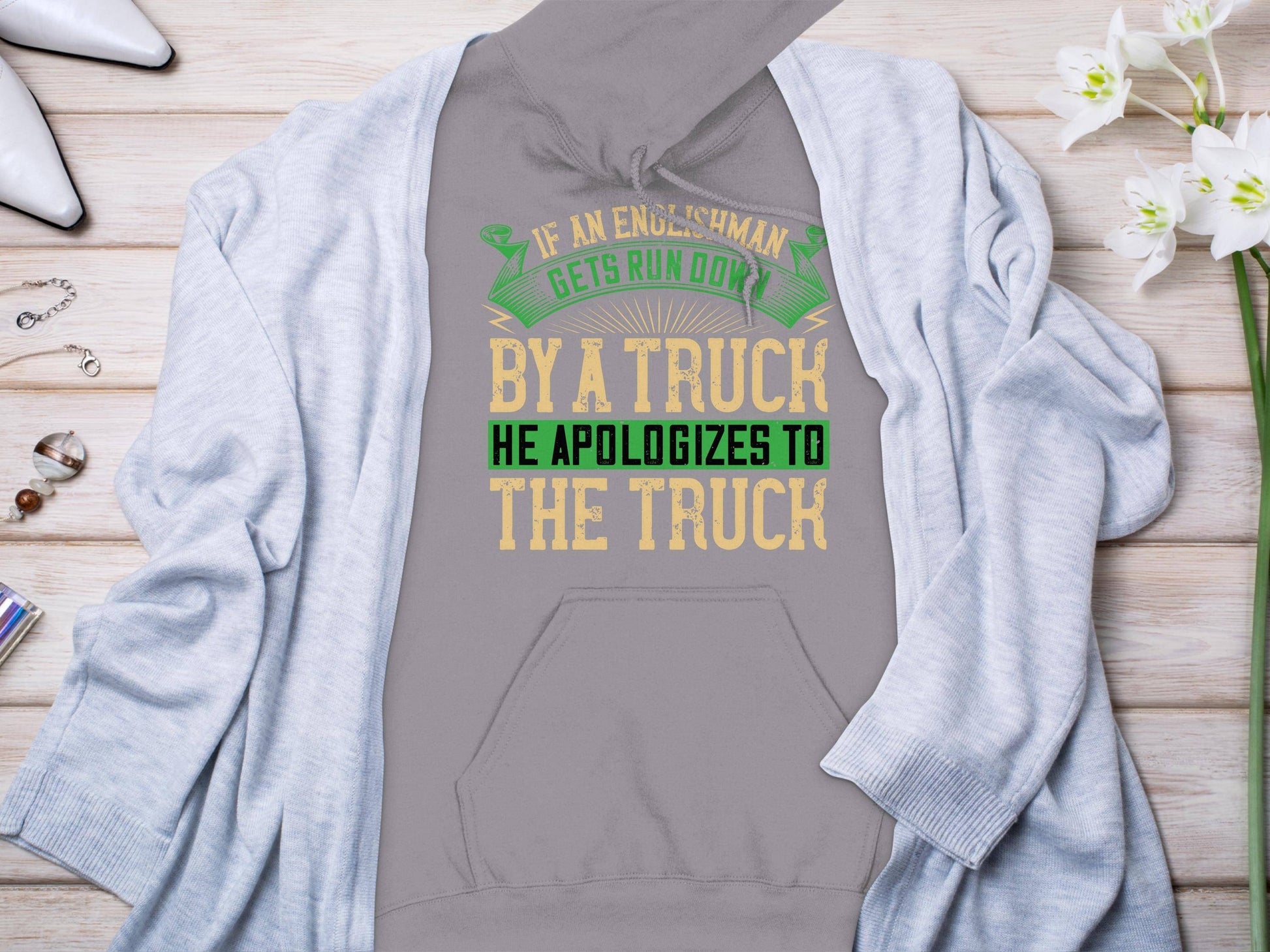 A gray hoodie from Garment Graphics features a humorous quote: If an Englishman gets run over by a truck, he apologizes to the truck in green, yellow, and white letters. The funny design is displayed under a light gray cardigan with decorations, capturing witty English etiquette.