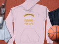 A light pink fishing hoodie by Garment Graphics, featuring Father and Son: Fishing Partners for Life, rests on a reddish surface, accompanied by a water bottle, towel, black shorts, and an orange basketball.