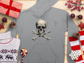 A Garment Graphics gray Skull and Crossbones Hoodie is set on wood, flanked by wrapped gifts, a white reindeer-patterned sweater, pine cones, a red and white lollipop, and festive red and white ornaments. Ideal for streetwear enthusiasts craving stylish comfort.