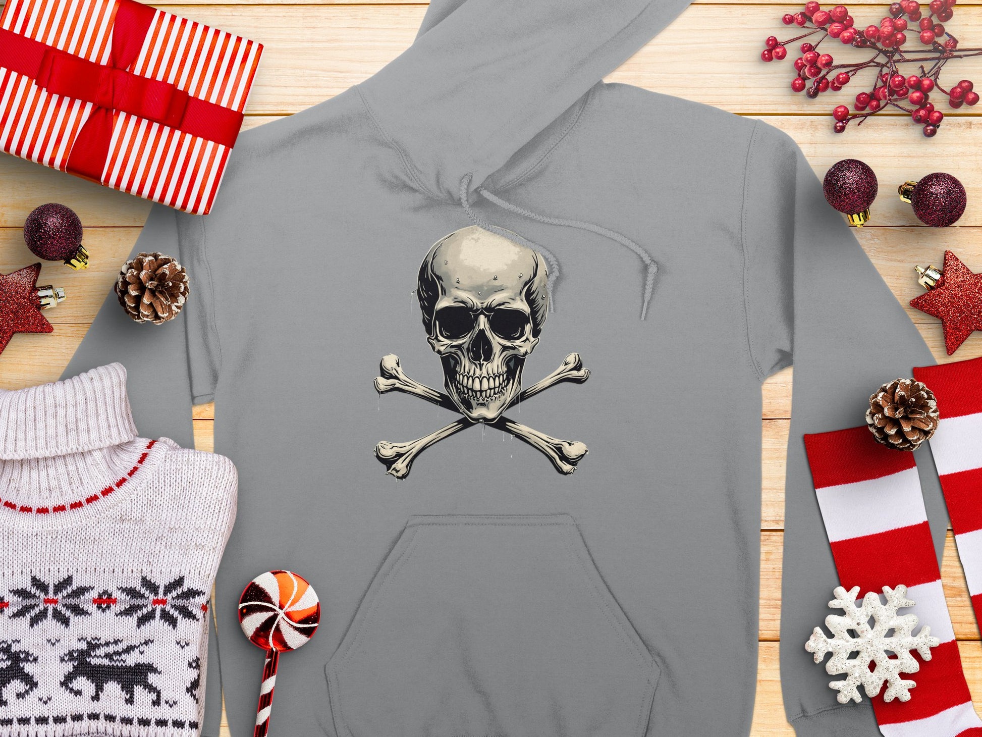 A Garment Graphics gray Skull and Crossbones Hoodie is set on wood, flanked by wrapped gifts, a white reindeer-patterned sweater, pine cones, a red and white lollipop, and festive red and white ornaments. Ideal for streetwear enthusiasts craving stylish comfort.