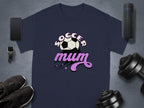 A navy graphic T-shirt by Garment Graphics, showcasing Soccer Mum in white and pink with a soccer ball, lies alongside a smartwatch, smartphone, wireless speaker, sneakers, and dumbbells on a gray surface.