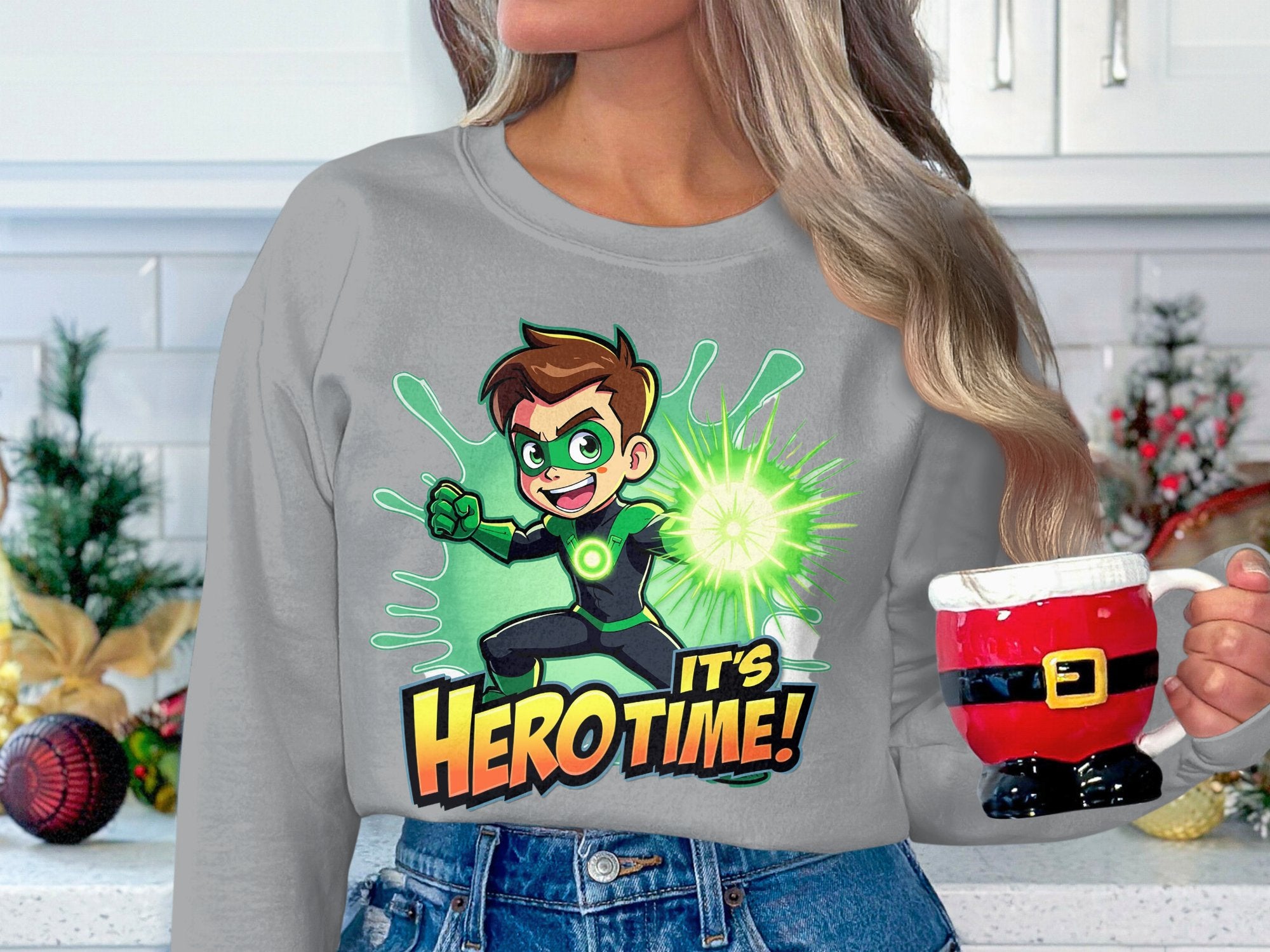 In a festive kitchen, someone with long hair holds a Santa-themed mug and wears Garment Graphics unisex Its Hero Time! sweatshirt, showcasing a green cartoon superhero. Holiday decorations, including ornaments and berries, are visible in the background.