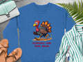 Garment Graphics presents a blue unisex sweatshirt featuring a playful cartoon turkey sprinting with the caption Turkey on the Run. Ideal for any occasion, it pairs well with tan sandals, a green purse, and a striped shirt against fern leaf accents.