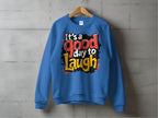 A Garment Graphics unisex blue sweatshirt hangs on a wooden hanger against a gray wall, showcasing colorful text, Its a good day to laugh, in a paint-splatter design on medium-heavy fabric.