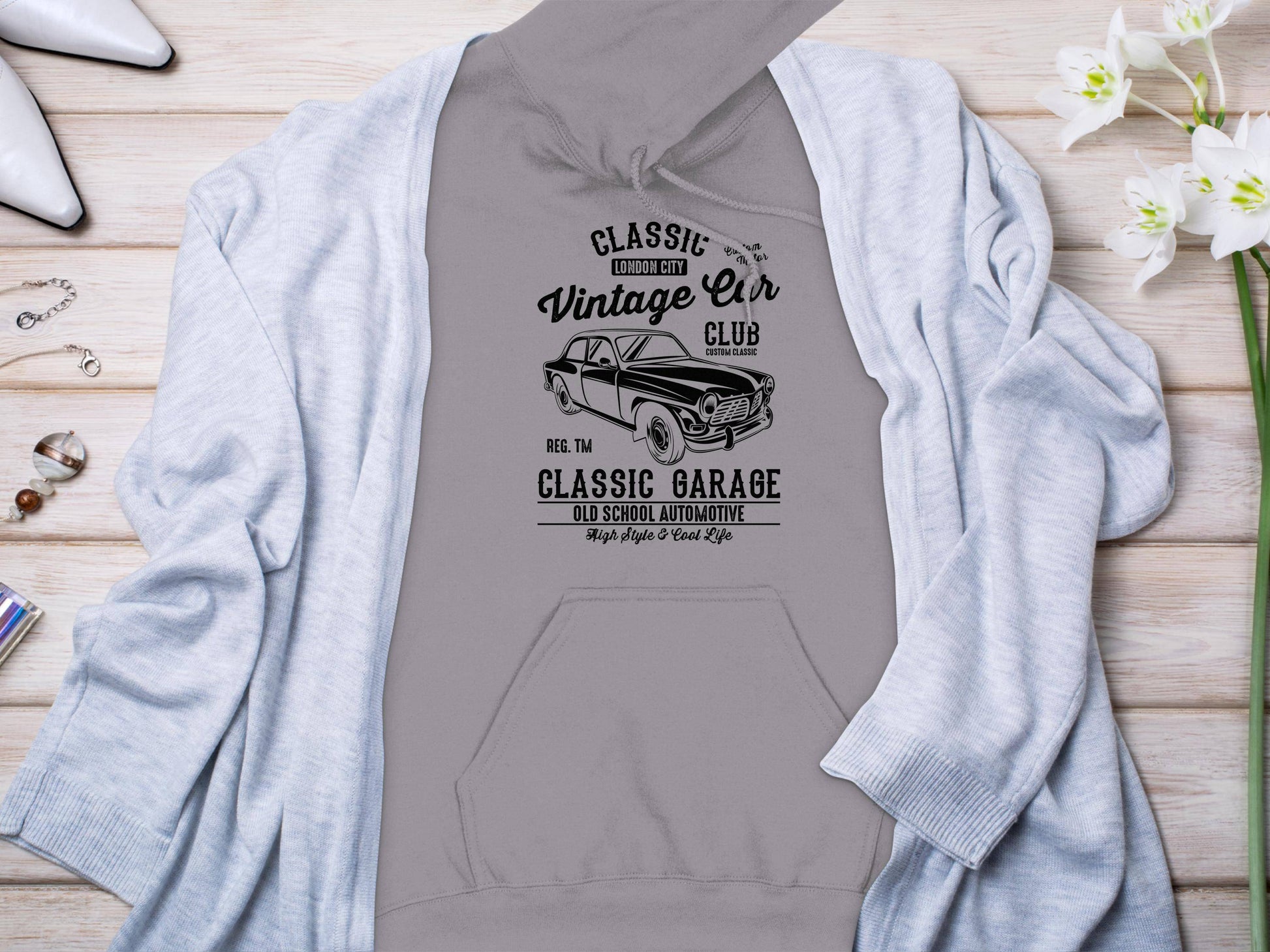 A gray hoodie by Garment Graphics with a Classic Car Vintage Car Club design lies on wood, partially under a light gray cardigan. White flowers, two lipsticks, a ring, and a silver bracelet complete the classic look for car enthusiasts.