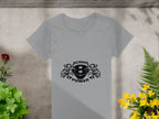 A gray T-shirt by Garment Graphics displays a central black design with SCANIA 8 POWER amid stylized flames or eagle heads. It is placed flat on concrete, decorated with surrounding plants and flowers.