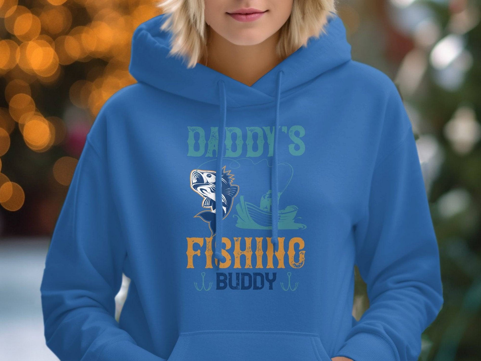 A person is wearing a blue hoodie from Garment Graphics Graphic Hoodies collection, featuring Daddys Fishing Buddy with fish and boat graphics. Softly blurred lights in the background suggest an ideal evening for outdoor adventures.