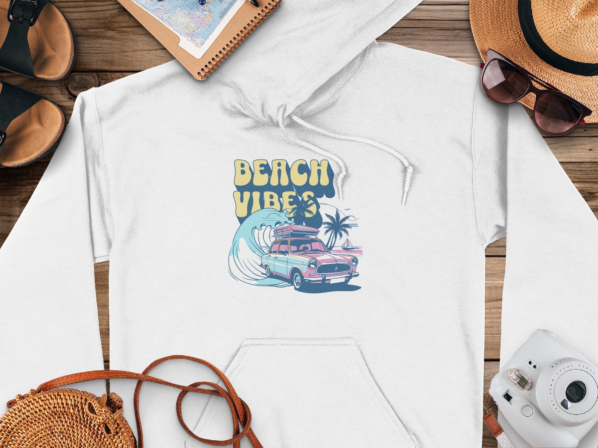 This white Garment Graphics hoodie, featuring a surfboard design with a pink vintage car, palm tree, ocean waves, and BEACH VIBES in yellow, exudes retro beach style. Positioned on wood alongside a camera, sandals, hat, and notebook, it captures vibrant coastal charm.