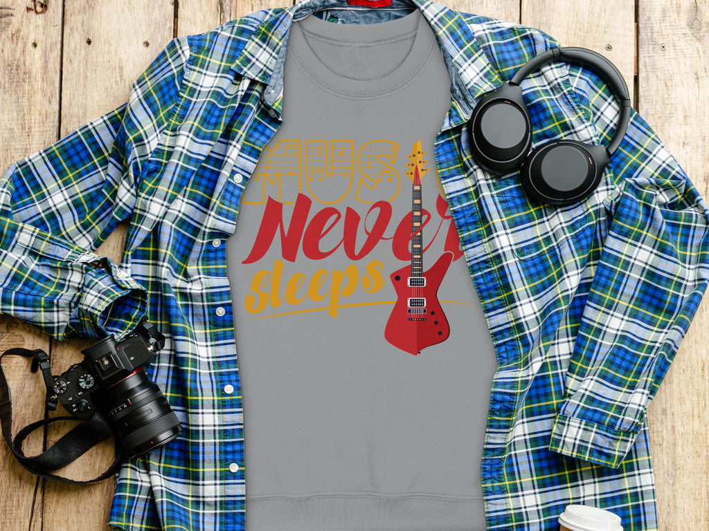 On a wooden surface, the Garment Graphics vibrant gray t-shirt features a red electric guitar and Music Never Sleeps. Ideal for music lovers, it pairs well with the blue and green plaid shirt, headphones, camera, and white cup.